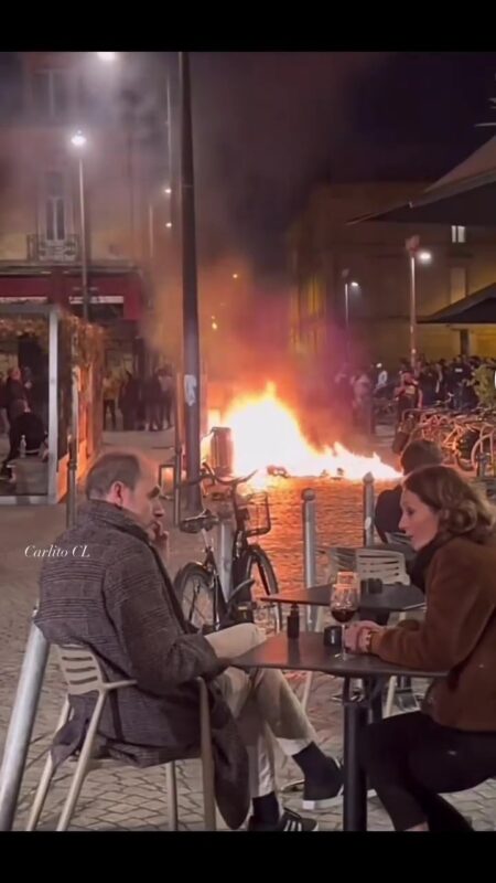 French couple enjoy glass of red wine as street burns behind them