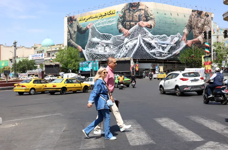 epa12918541 Iranians walk past a huge billboard carrying a sentence reading in Persian 'The Strait of Hormuz remains closed' at Enghelab Square in Tehran, <a href=